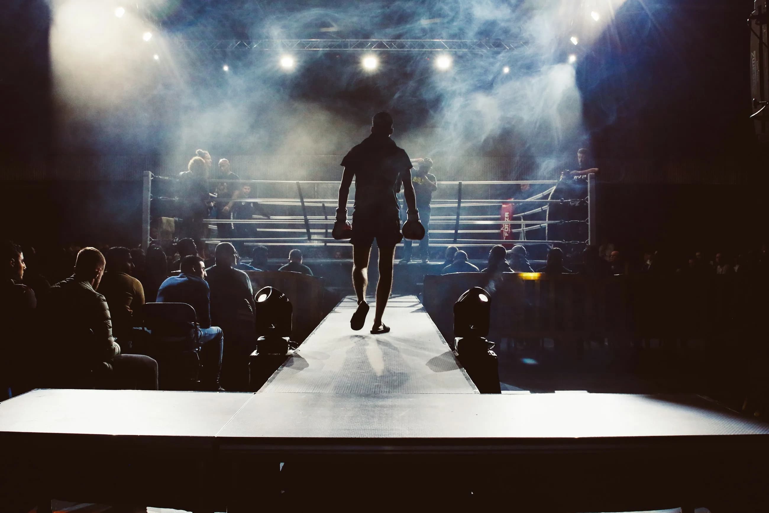 A fighter seen from behind walking out to a lit ring through smoke